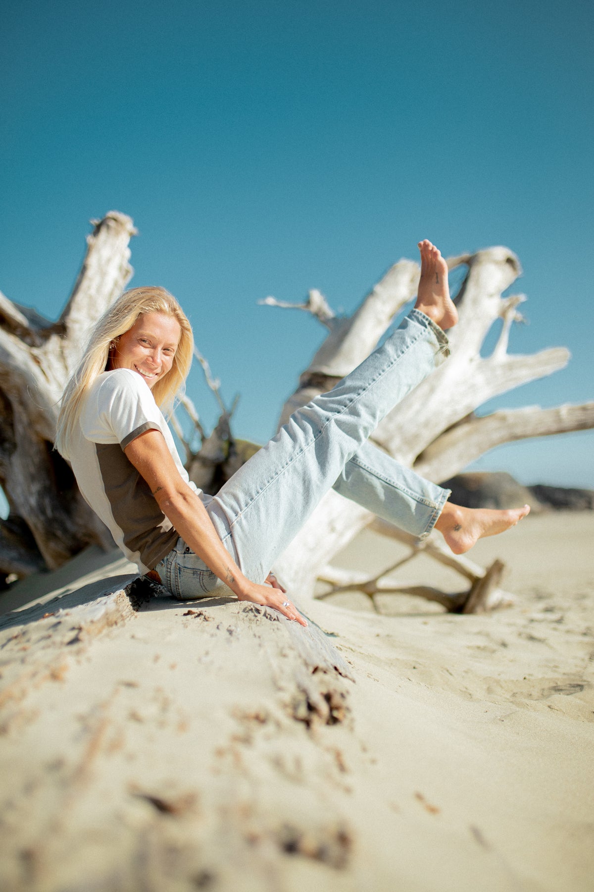 A woman sitting on a sandy beach with driftwood behind her, wearing a white t-shirt, brown vest, and light blue jeans against a clear blue sky.