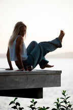 Cougar Milk | A woman sits barefoot on the edge of a wooden dock over calm water at sunset, wearing a white sleeveless top and loose blue jeans, with a glass of red wine beside her.