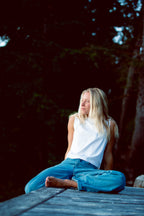 Cougar Milk | A woman with blonde hair sits cross-legged on a weathered wooden dock in a dark, wooded outdoor setting. She is wearing a white sleeveless top and blue jeans.