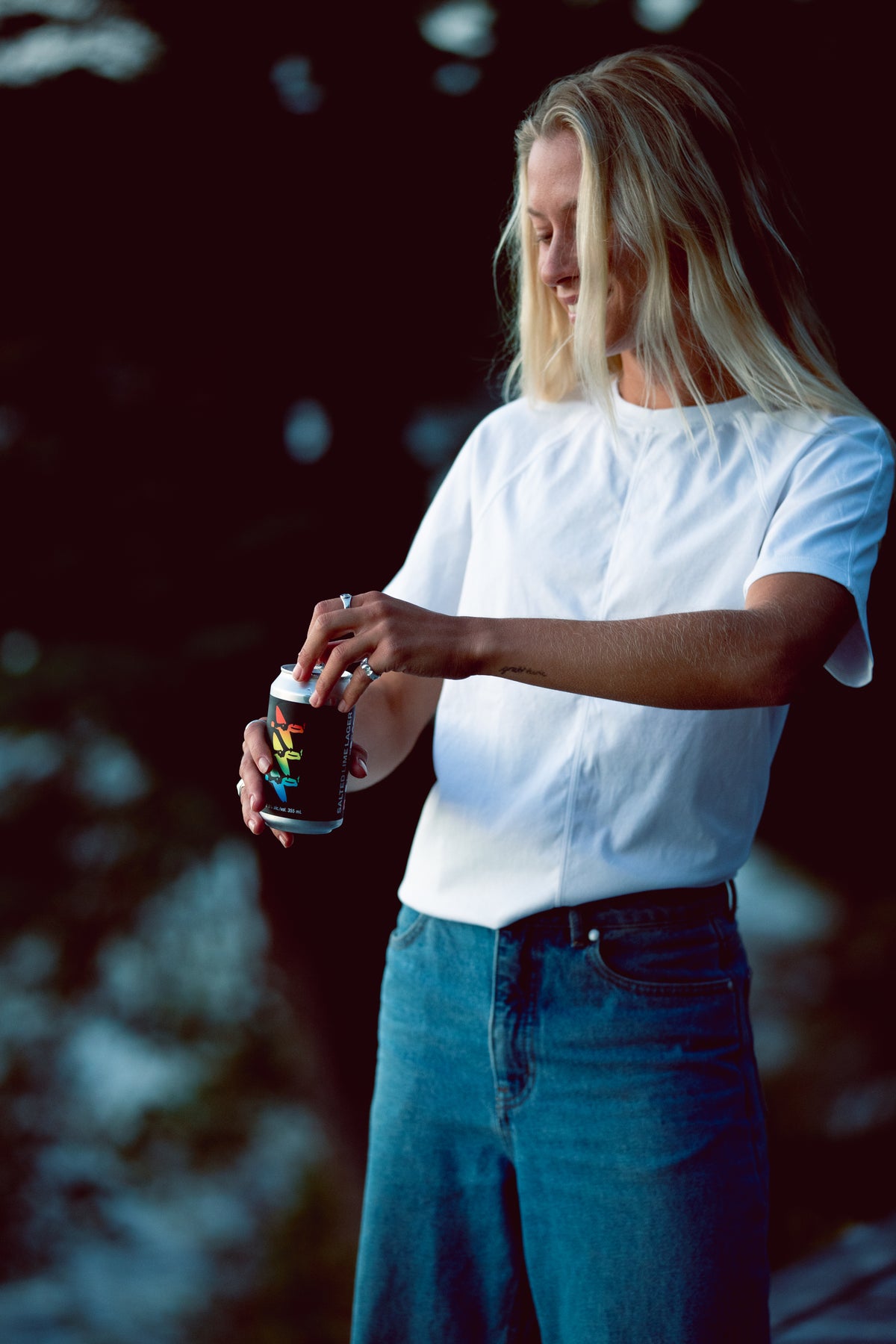 A woman with blonde hair wearing a white short-sleeve t-shirt and blue jeans stands outdoors, holding a black can with a colorful graphic label.