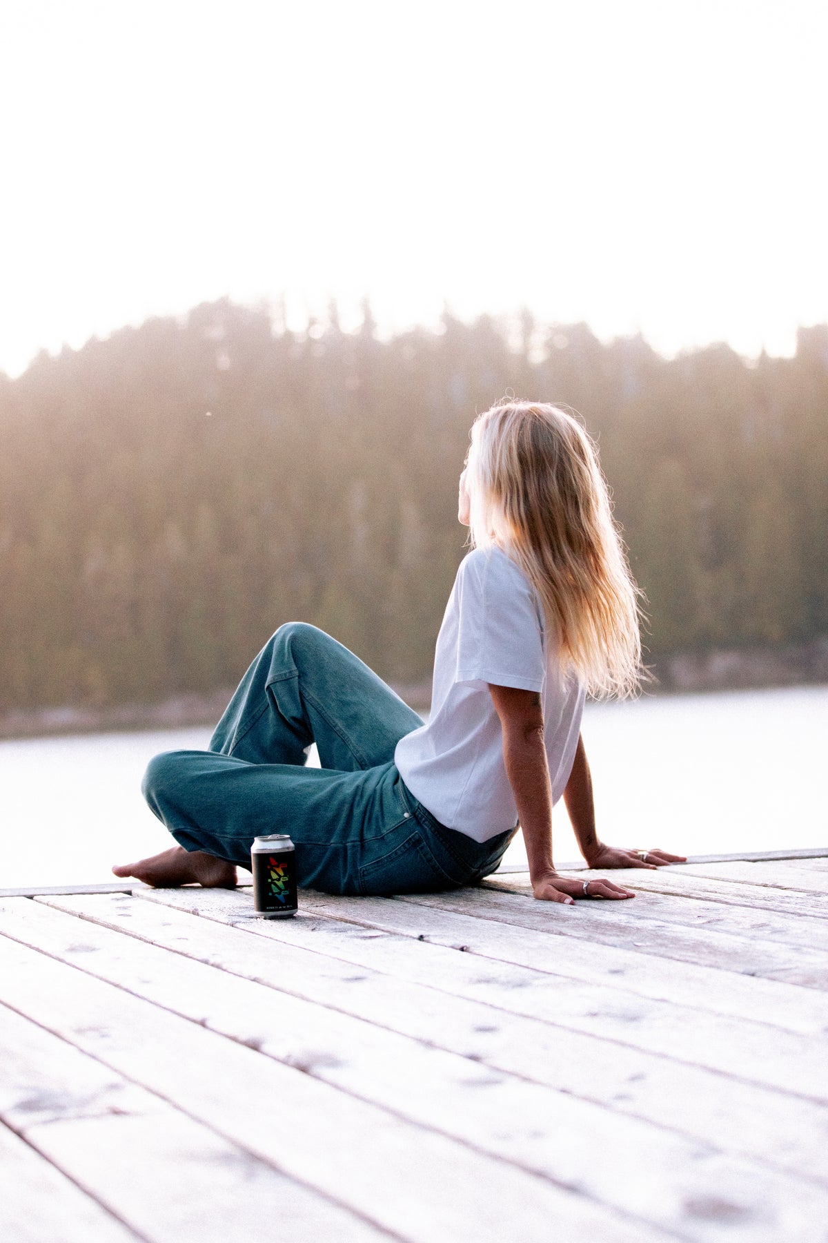 A woman with long blonde hair sits on a wooden dock by a lake, seen from behind, wearing a white t-shirt and blue jeans. A dark beverage can sits beside her, with a blurred forest background under a bright sky.
