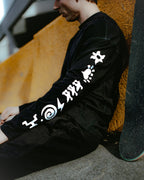 Ink Black | Lifestyle Skate A person wearing a black long-sleeve shirt with white graphic symbols printed down the sleeves, sitting against a textured orange wall.
