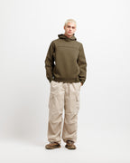 Olive Pit | On Model Full Front A model stands against a light gray studio background wearing an olive green hooded sweatshirt, beige cargo pants, and brown shoes.