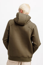 Olive Pit | On Model Back A person viewed from behind wearing an olive green pullover hoodie with a high collar and zippered pockets, paired with light beige pants, against a plain white background.