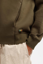 Olive Pit | On Model Detail Close-up of a person wearing an olive green zip-up hoodie with a small colorful embroidered logo patch near the pocket, paired with light beige pants against a plain white background.