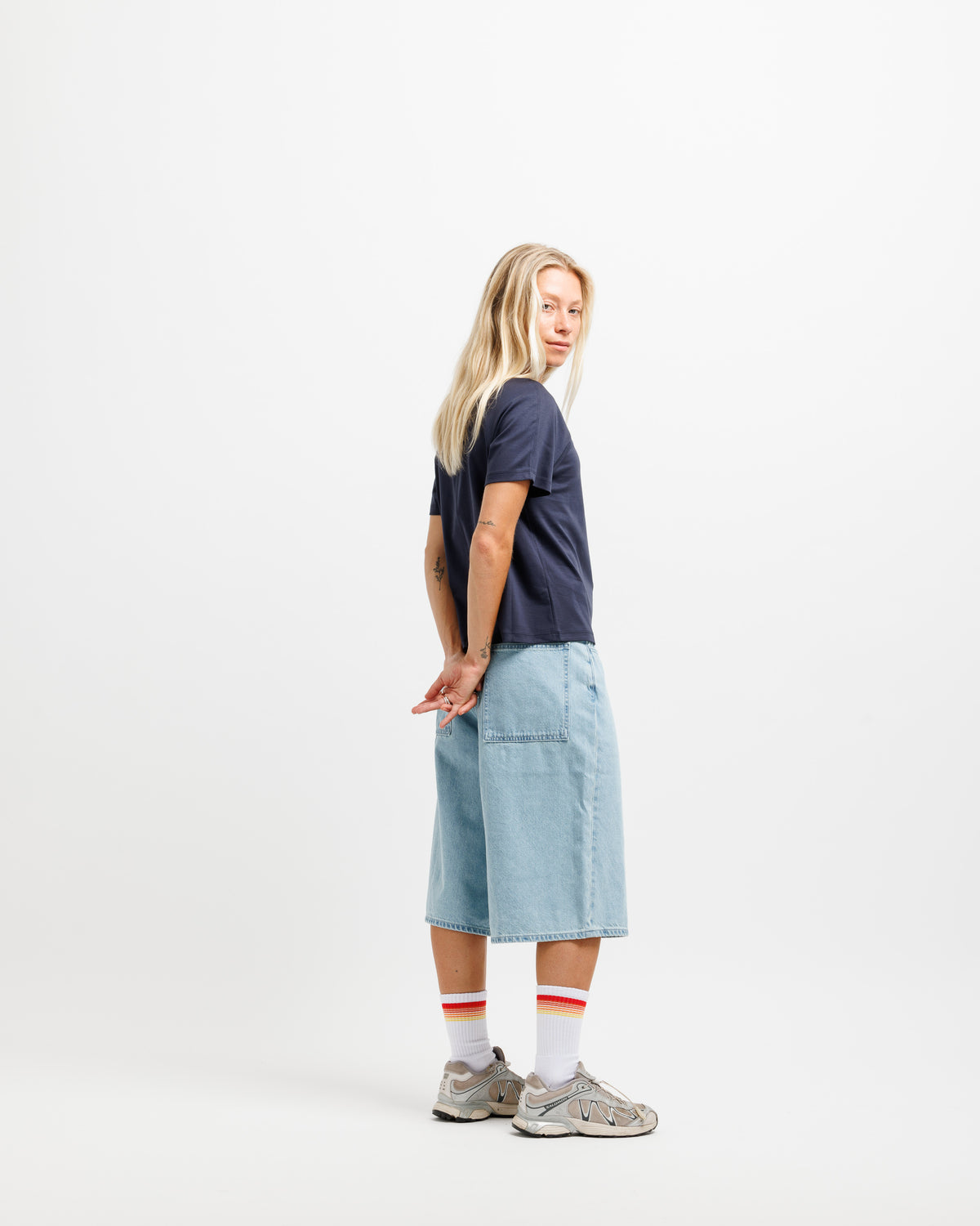 A woman stands in profile against a plain white studio background, wearing a dark navy short-sleeve T-shirt, light blue denim shorts, white socks with red stripes, and gray sneakers.