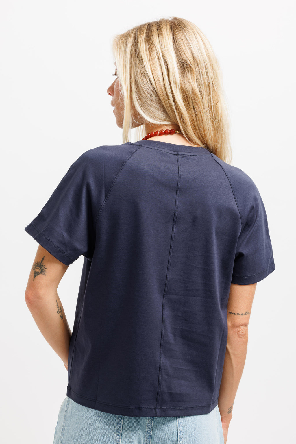 A woman viewed from behind wearing a navy blue short-sleeve t-shirt with a central seam, paired with light blue jeans, against a plain white background.