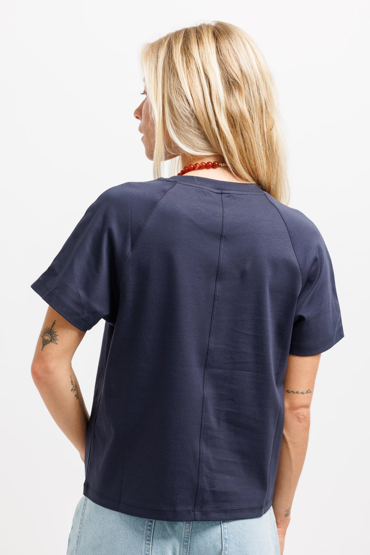 A woman viewed from behind wearing a navy blue short-sleeve t-shirt with a central seam, paired with light blue jeans, against a plain white background.