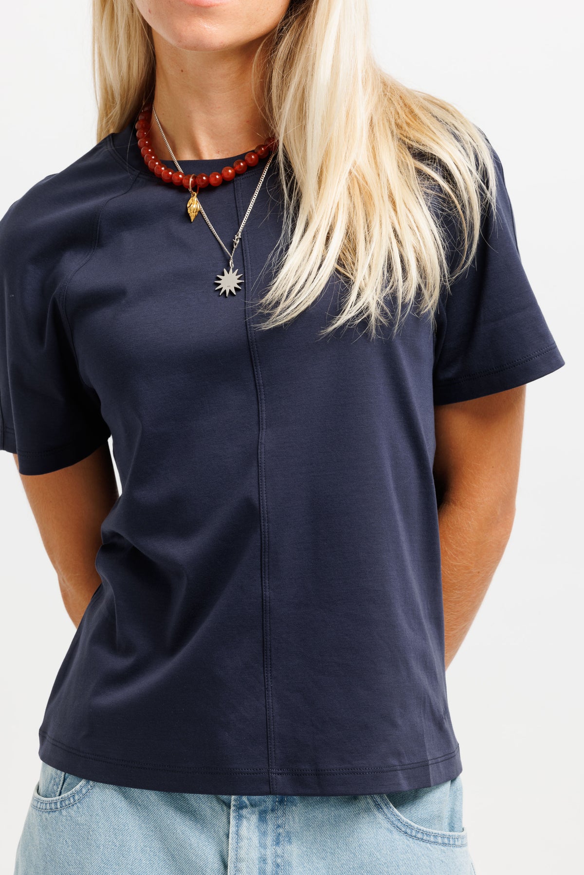 A woman wearing a navy blue short-sleeve t-shirt with a front zipper, paired with light blue jeans and layered necklaces, shown against a plain white background.