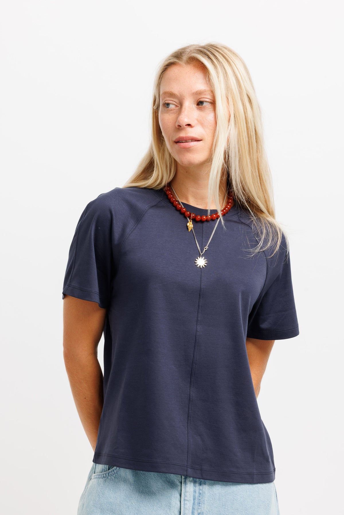 A woman with blonde hair wearing a navy blue short-sleeve t-shirt and light blue jeans, accessorized with layered necklaces, photographed against a plain white background.