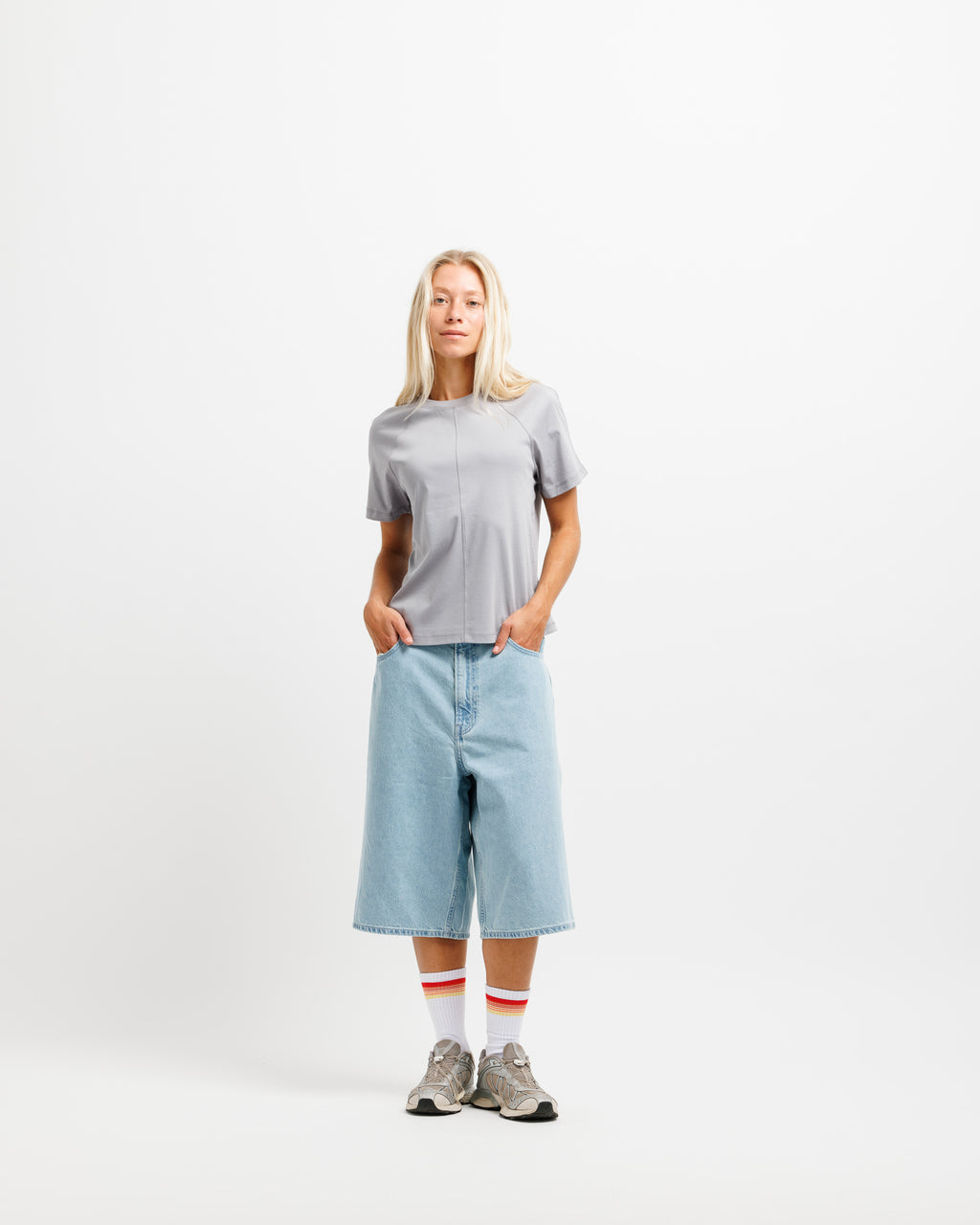 A woman stands against a plain white studio background wearing a light gray short-sleeve top, light blue cropped denim shorts, white socks with red stripes, and gray sneakers.