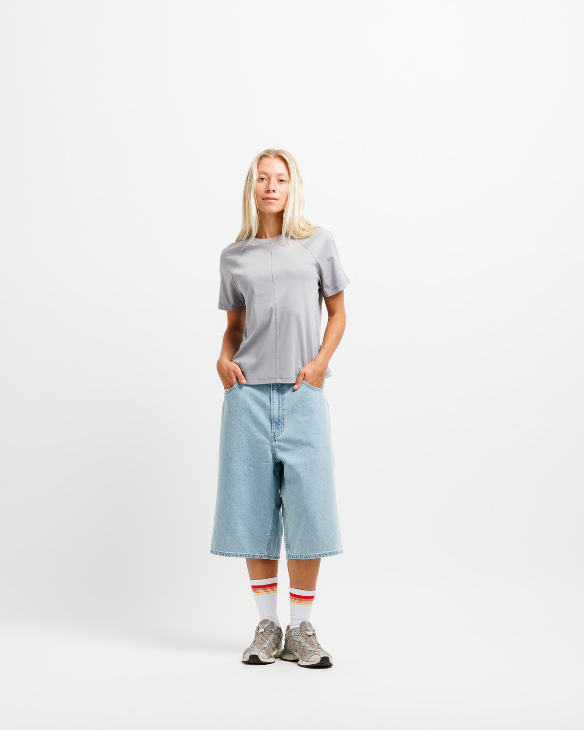 A woman stands against a plain white studio background wearing a light gray short-sleeve top, light blue cropped denim shorts, white socks with red stripes, and gray sneakers.