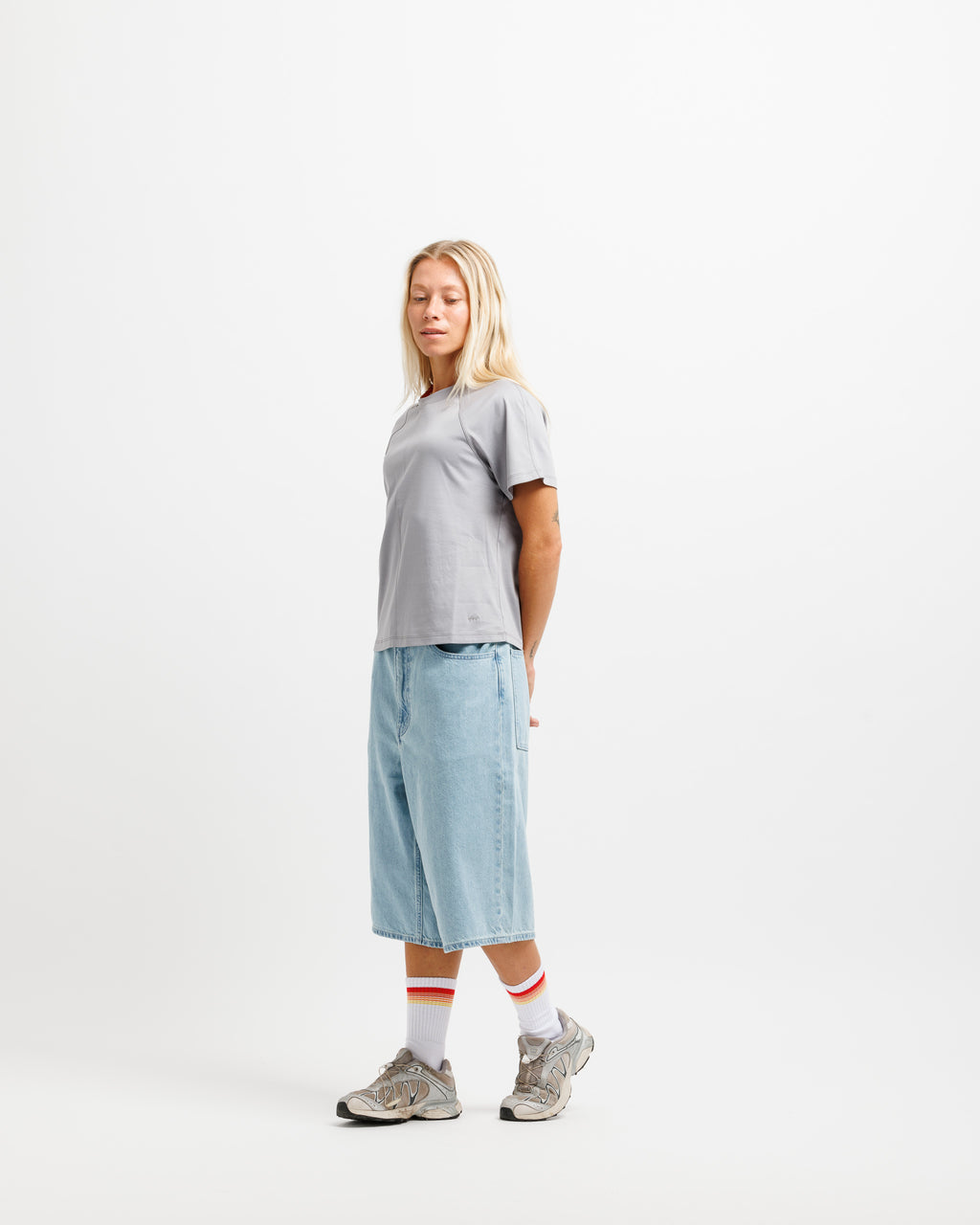 A woman stands in profile wearing a light gray short-sleeve t-shirt, light blue denim shorts, white crew socks with red stripes, and gray sneakers, against a plain white studio background.