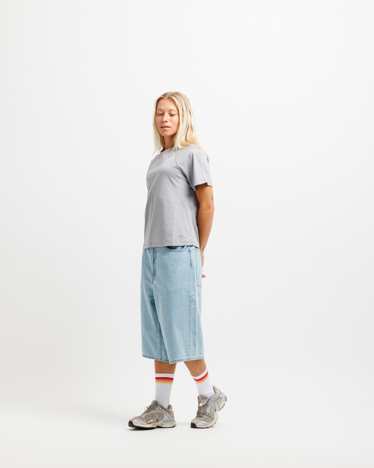 A woman stands in profile wearing a light gray short-sleeve t-shirt, light blue denim shorts, white crew socks with red stripes, and gray sneakers, against a plain white studio background.