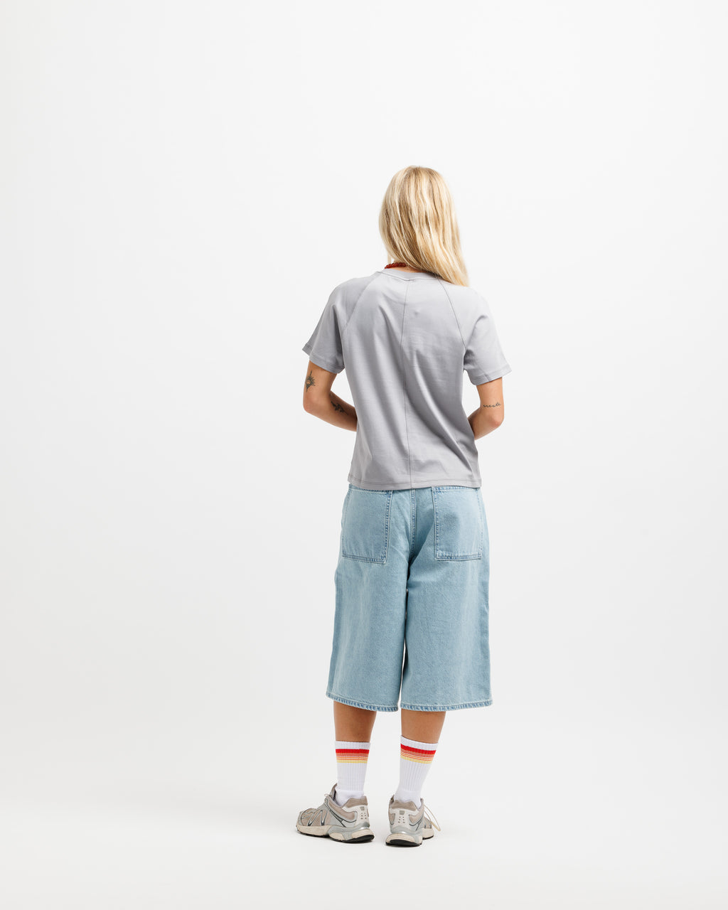 A model viewed from behind wearing a light gray t-shirt, light blue denim shorts, white socks with red stripes, and gray sneakers, set against a plain white studio background.