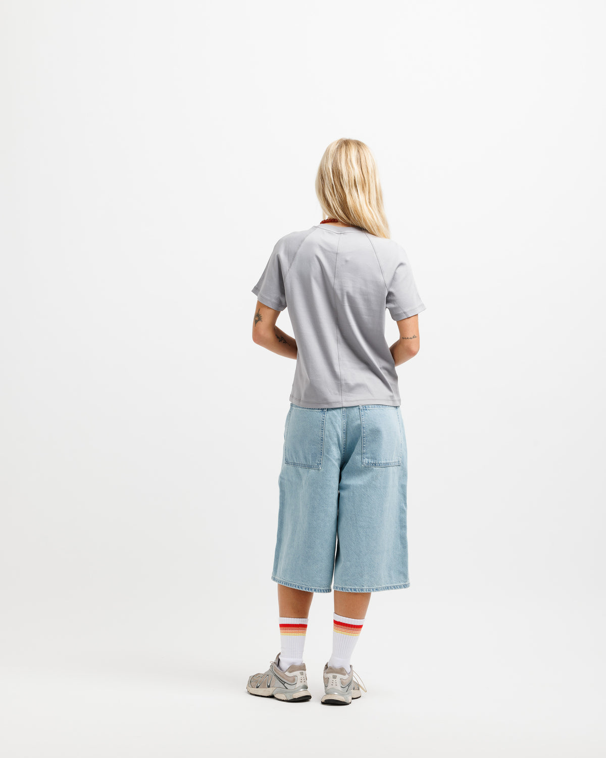 A model viewed from behind wearing a light gray t-shirt, light blue denim shorts, white socks with red stripes, and gray sneakers, set against a plain white studio background.