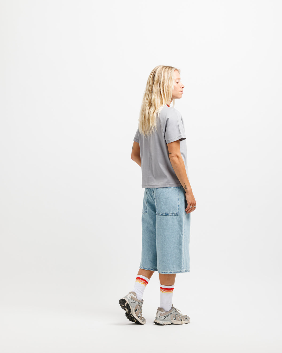 A woman stands in profile against a clean white studio background, wearing a light gray short-sleeve T-shirt, light blue denim shorts, white socks with orange stripes, and gray sneakers.