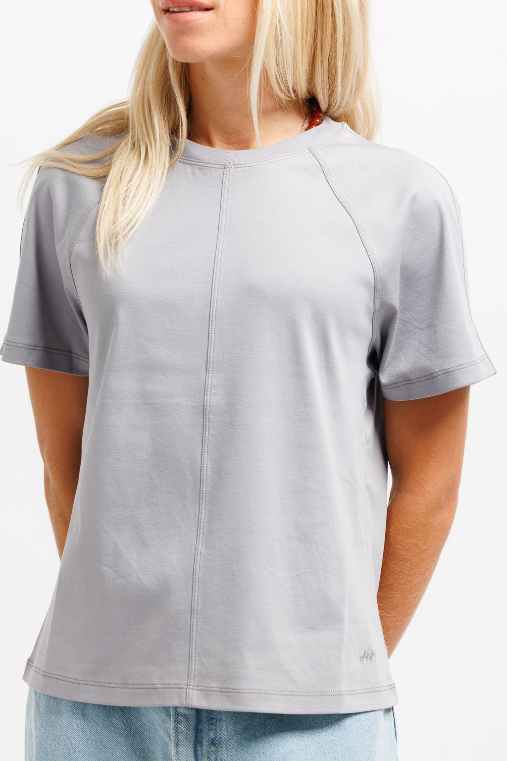 A woman wearing a light gray short-sleeve crewneck t-shirt with a vertical seam down the center, paired with light blue jeans against a plain white background.