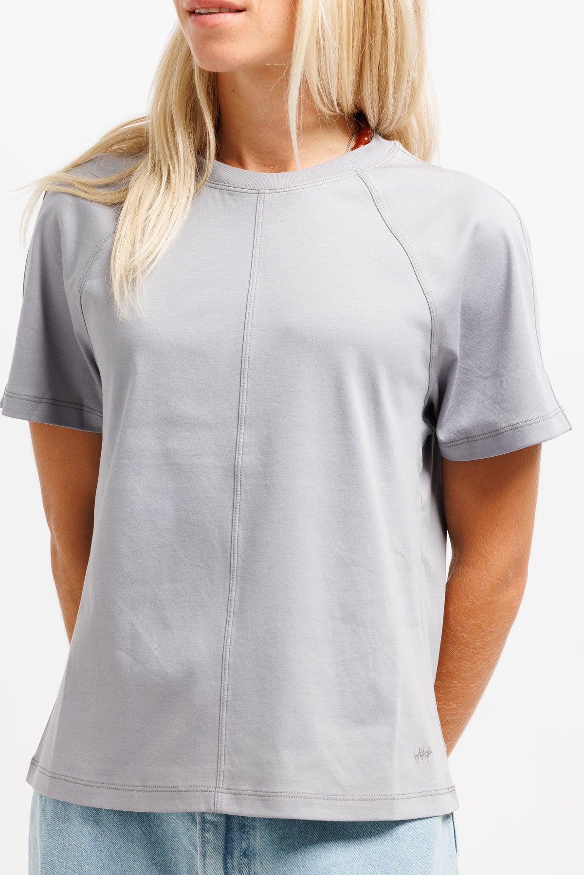 A woman wearing a light gray short-sleeve crewneck t-shirt with a vertical seam down the center, paired with light blue jeans against a plain white background.