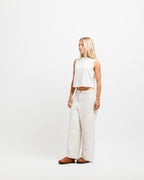 Cougar Milk | A blonde model stands in profile wearing a white sleeveless crop top and off-white wide-leg pants with brown leather shoes, photographed against a seamless white studio background.