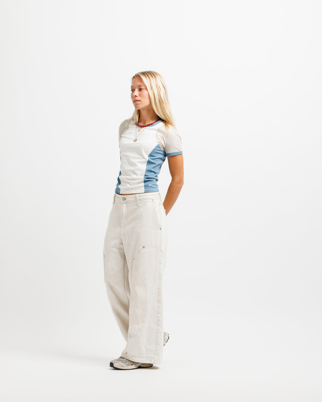 A blonde woman stands in profile wearing a fitted short-sleeve top with white and light blue color-blocked panels, paired with wide-leg off-white pants and gray sneakers, against a plain white studio background.