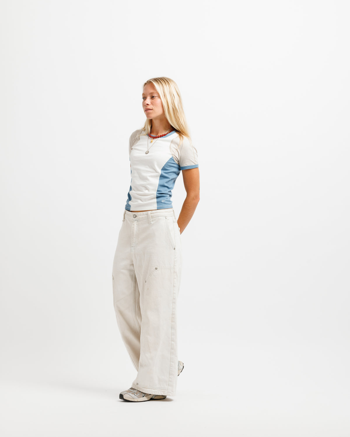 A blonde woman stands in profile wearing a fitted short-sleeve top with white and light blue color-blocked panels, paired with wide-leg off-white pants and gray sneakers, against a plain white studio background.