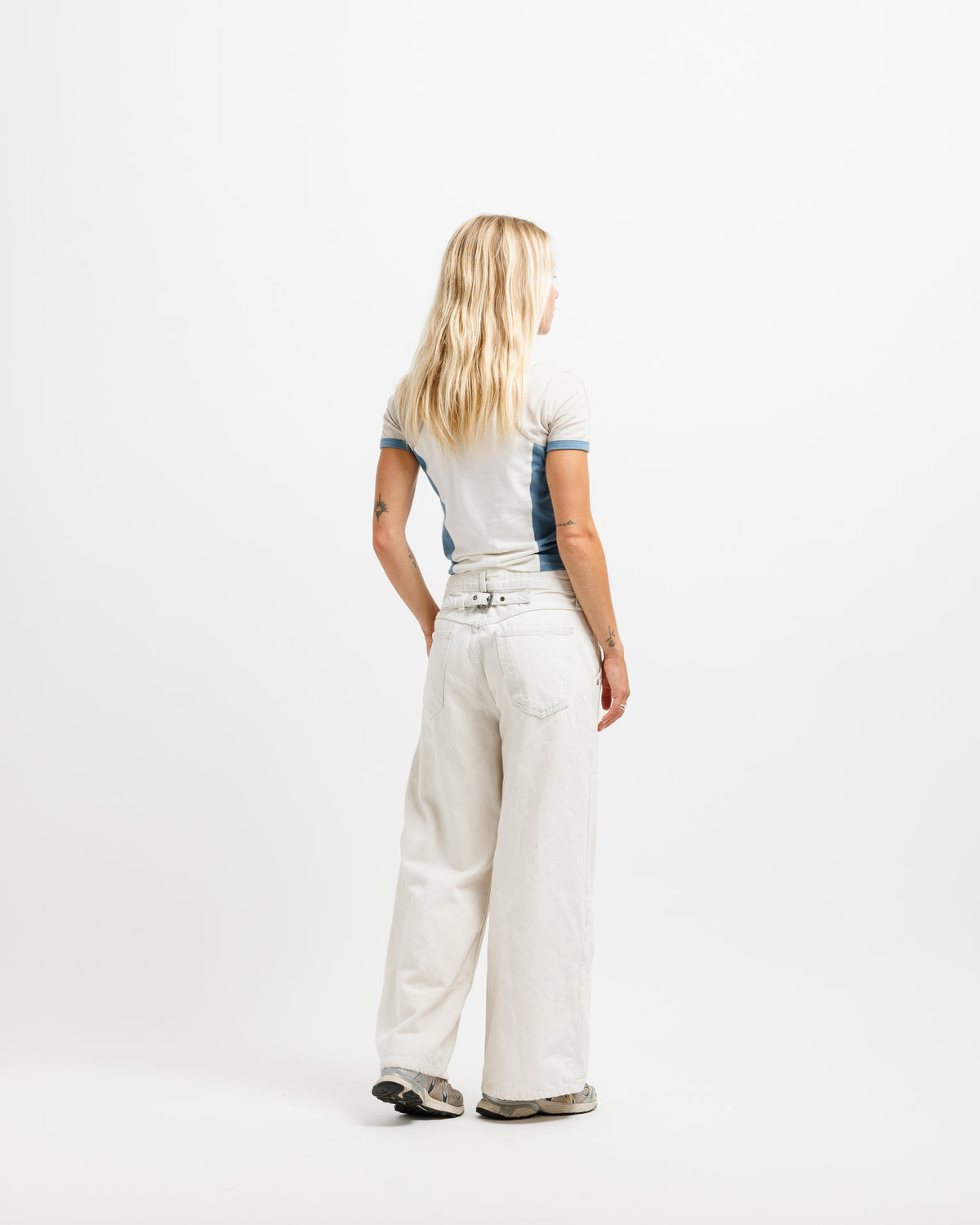 A woman with long blonde hair is shown from behind wearing a light gray t-shirt with blue side panels and off-white wide-leg jeans, standing against a solid white studio background.