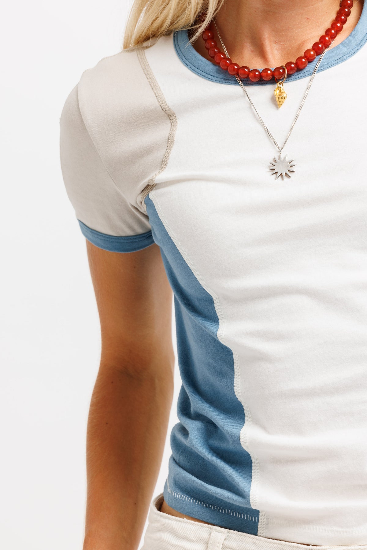 A close-up of a person wearing a short-sleeve ringer T-shirt with a white body, light blue collar and sleeve trim, and a vertical light blue panel on the side. The person is also wearing layered necklaces, including a red beaded necklace and a silver sunburst pendant.