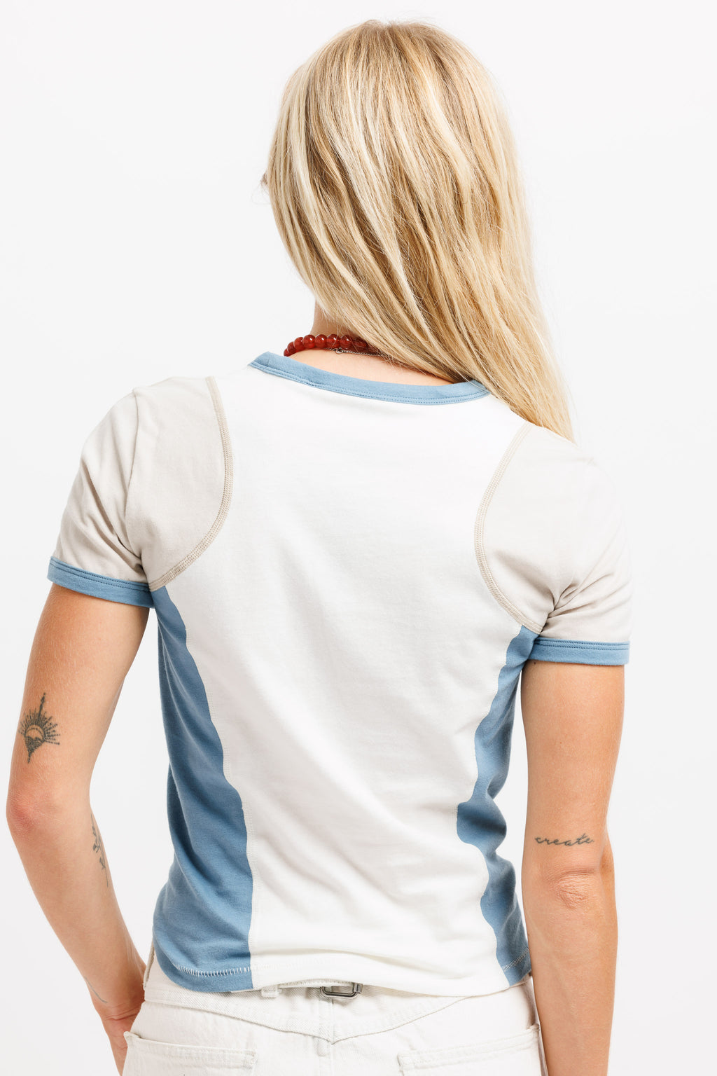 A woman viewed from behind wearing a white ringer T-shirt with light blue trim on the collar, sleeve cuffs, and side panels, paired with white pants against a plain white studio background.