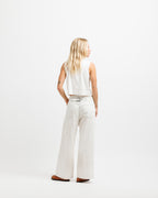Cougar Milk | A woman with blonde hair is shown from behind wearing a white sleeveless crop top and matching white wide-leg trousers with a silver belt, standing against a solid white studio background.