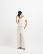 Cougar Milk | On Model Full Front A woman stands against a plain white studio background wearing a navy baseball cap with white '79' lettering, a white short-sleeve T-shirt, off-white wide-leg pants, and gray sneakers.