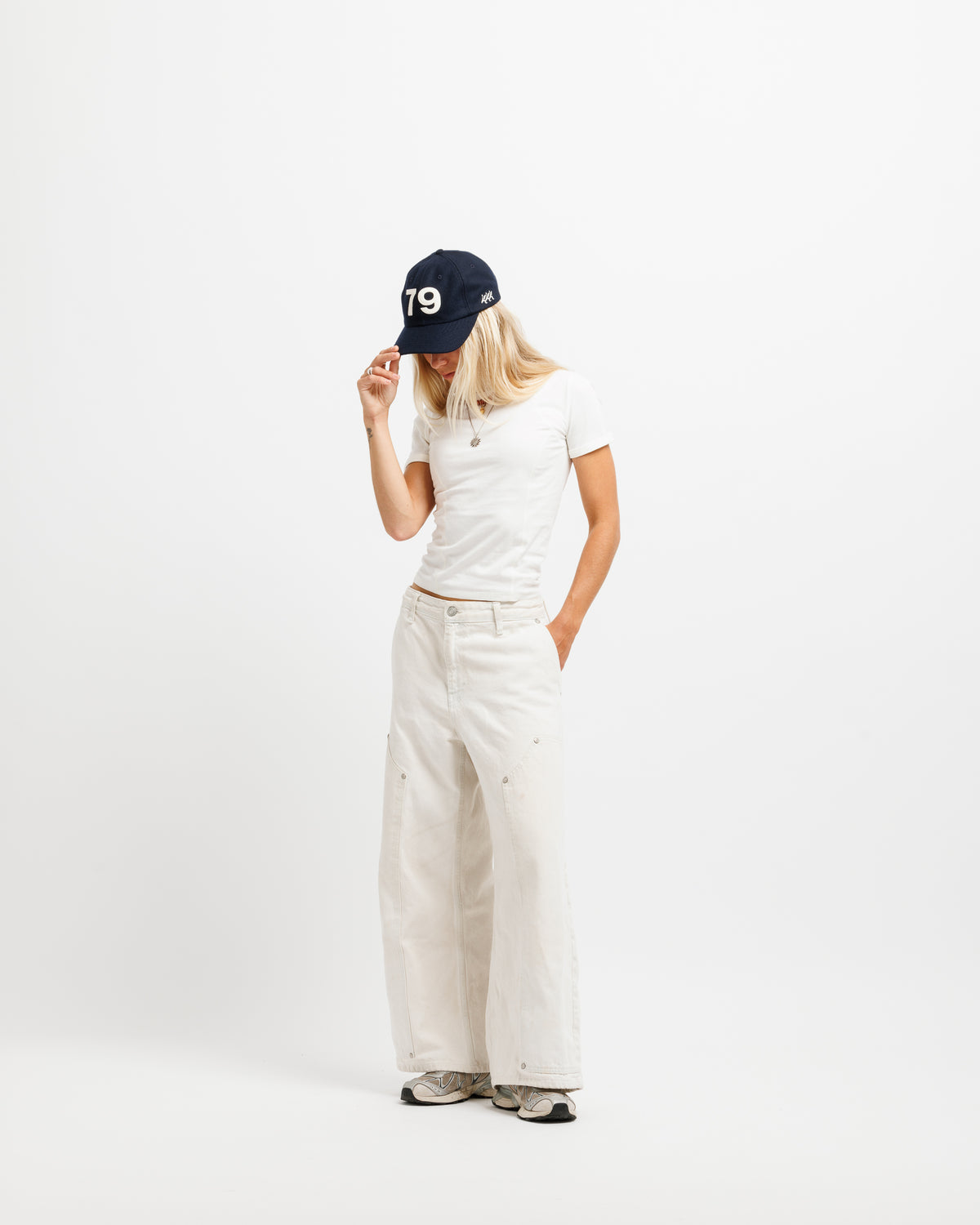 A woman stands against a plain white studio background wearing a navy baseball cap with white '79' lettering, a white short-sleeve T-shirt, off-white wide-leg pants, and gray sneakers.