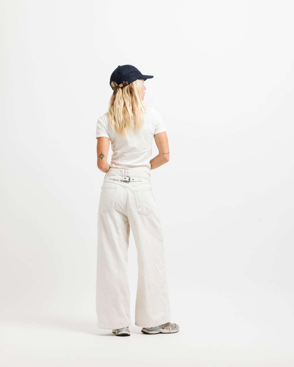 A woman viewed from behind wearing a navy blue baseball cap, a white t-shirt, off-white wide-leg pants with a matching belt, and gray sneakers, standing against a solid white studio background.