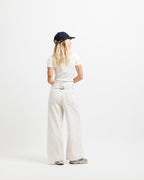 Cougar Milk | On Model Full Back A woman viewed from behind wearing a navy blue baseball cap, a white t-shirt, off-white wide-leg pants with a matching belt, and gray sneakers, standing against a solid white studio background.