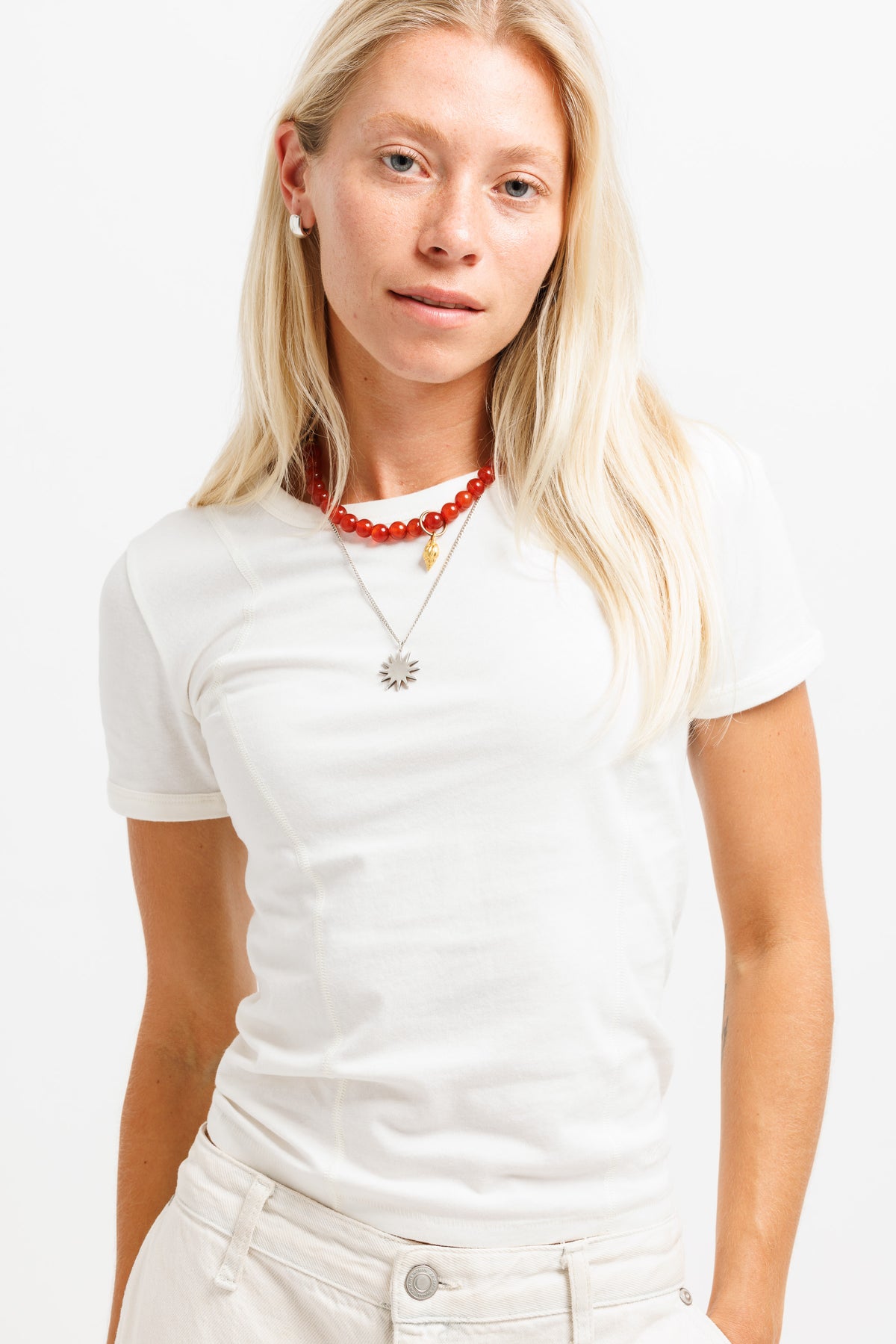 A blonde woman wearing a white short-sleeve crewneck t-shirt, white jeans, and layered necklaces, including a red beaded necklace and a silver sunburst pendant, against a plain white background.