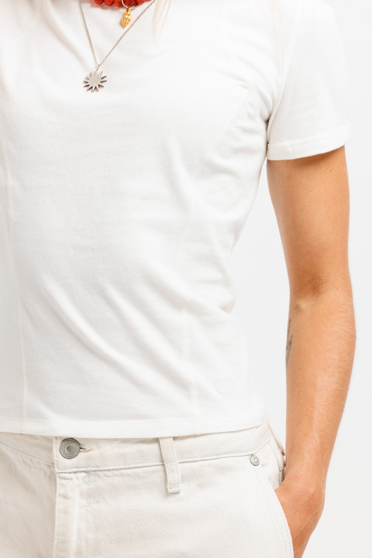 A close-up of a person wearing a fitted white short-sleeve crew-neck t-shirt and off-white jeans, accessorized with a silver sunburst pendant necklace.