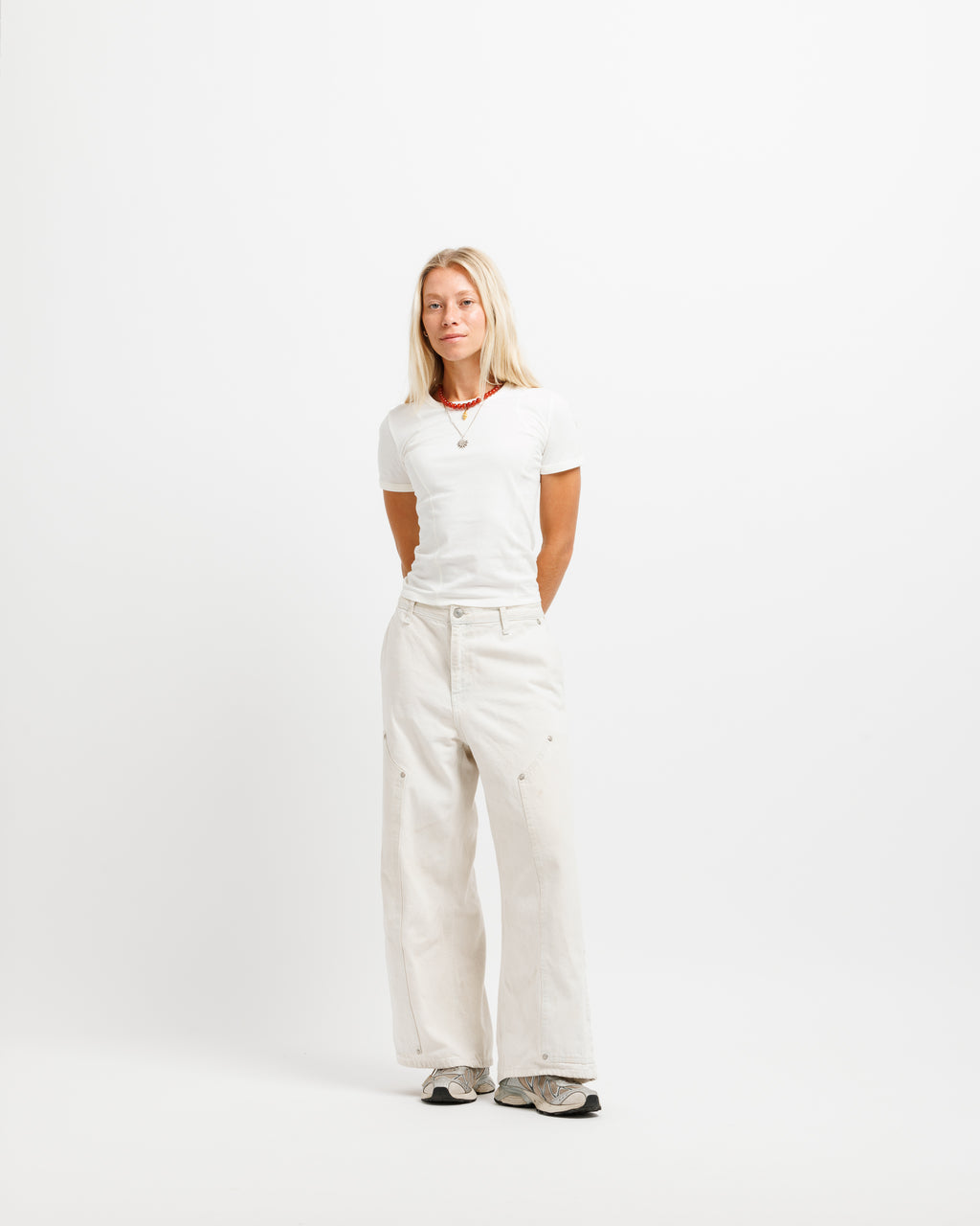 A blonde woman stands against a plain white studio background wearing a white short-sleeve t-shirt, off-white wide-leg trousers, and gray sneakers.