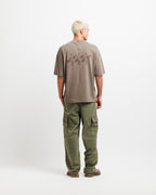 Cabin Smoke | Back A man viewed from the back wearing a taupe-brown short-sleeve t-shirt with a dark graphic on the back and olive green cargo pants, standing against a plain white studio background.