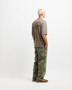 Cabin Smoke | A man with platinum blonde hair stands in profile facing away from the camera, wearing a taupe-brown t-shirt with a black graphic on the back and olive green cargo pants, against a plain white studio background.