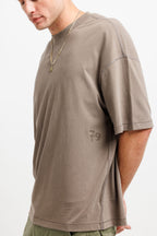 Cabin Smoke | A man wearing an oversized taupe-brown t-shirt with a tonal '79' logo on the lower left chest, paired with olive green pants and a gold chain necklace, photographed against a clean white background.