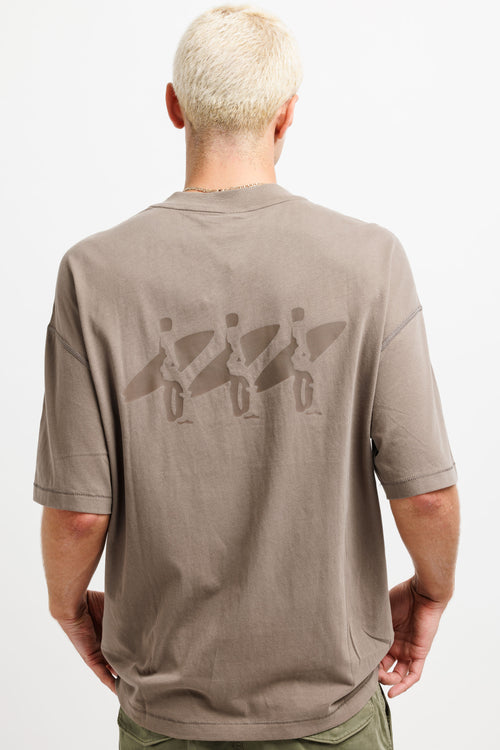 A man viewed from behind wearing a taupe brown t-shirt with a tonal graphic of three surfers carrying boards across the back, paired with olive green pants against a white studio background.