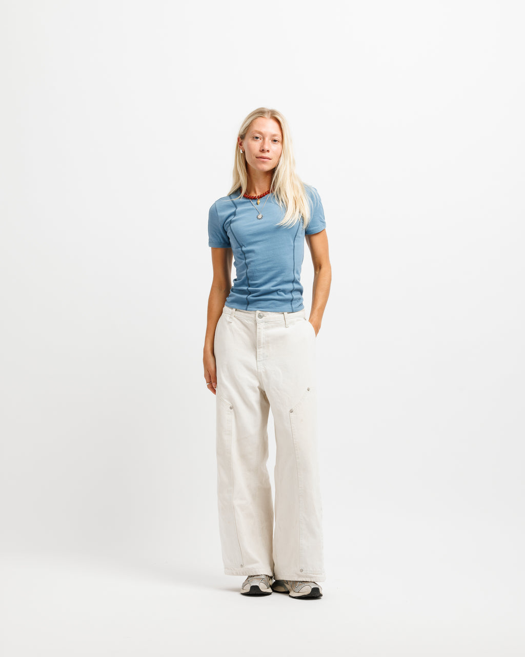 A blonde woman stands against a plain white studio background wearing a fitted blue short-sleeve top, off-white wide-leg pants, and gray sneakers.