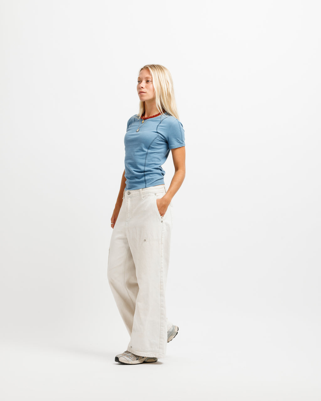 A blonde woman stands in profile wearing a fitted light blue short-sleeve top, off-white wide-leg trousers, and gray sneakers against a plain white studio background.