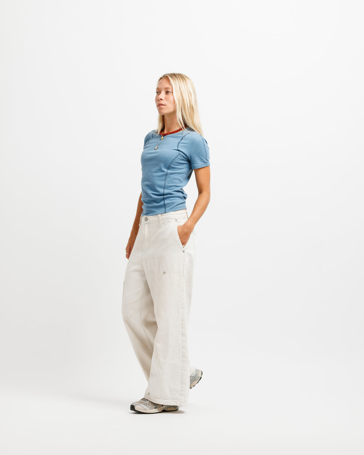 A blonde woman stands in profile wearing a fitted light blue short-sleeve top, off-white wide-leg trousers, and gray sneakers against a plain white studio background.