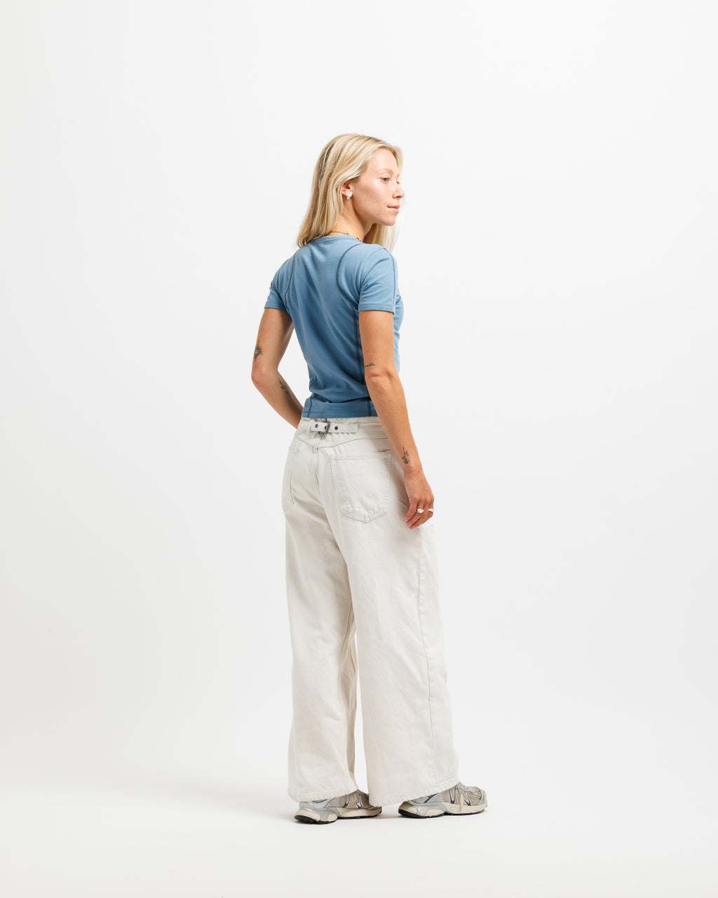 A woman with blonde hair is shown from a back-three-quarter view wearing a blue t-shirt, off-white wide-leg jeans, and gray sneakers against a plain white studio background.