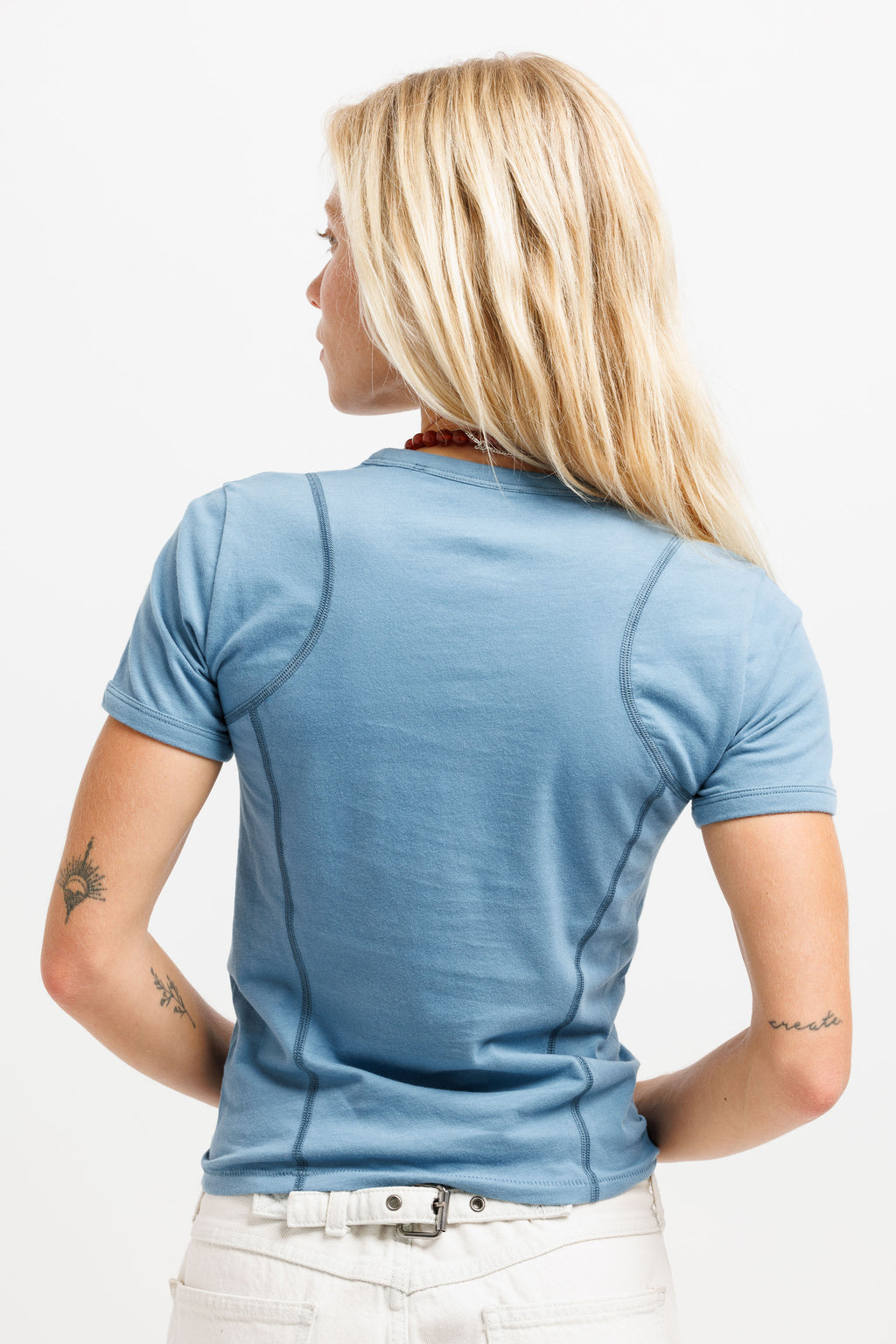 A woman with blonde hair is shown from the back wearing a light blue short-sleeve athletic t-shirt with contrast stitching, paired with white pants, against a plain white background.