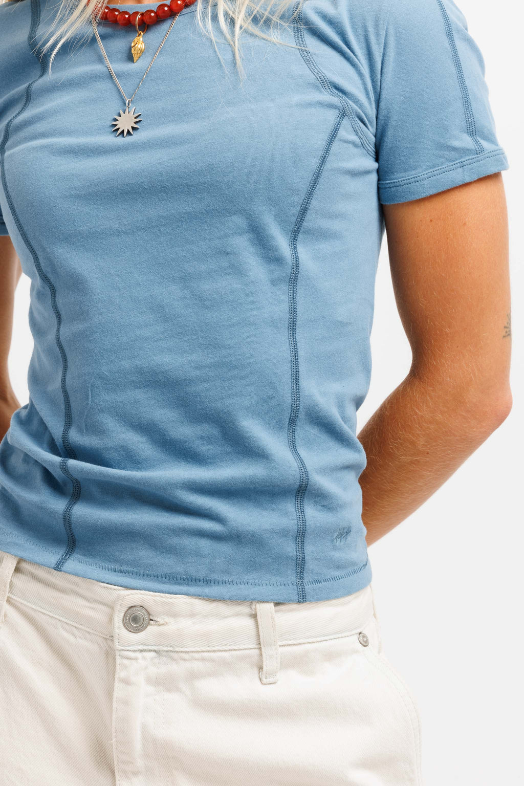 A close-up of a person wearing a light blue fitted short-sleeve t-shirt with visible seam detailing, paired with off-white pants. The person is also wearing layered necklaces, including a red beaded necklace and a gold sunburst pendant.