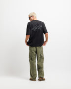 Ink Black | Back A man with short blonde hair and glasses, viewed from behind, wearing a black t-shirt with a dark westbeach graphic on the back, olive green cargo pants, and brown shoes, standing against a plain white studio background.
