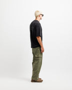 Ink Black | A man with blonde hair and sunglasses stands in profile wearing a black t-shirt, olive green cargo pants, and brown shoes against a plain white studio background.