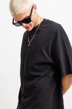 Ink Black | A man with short blonde hair wearing a black oversized t-shirt, black sunglasses, and a gold chain necklace, looking down against a plain white background.